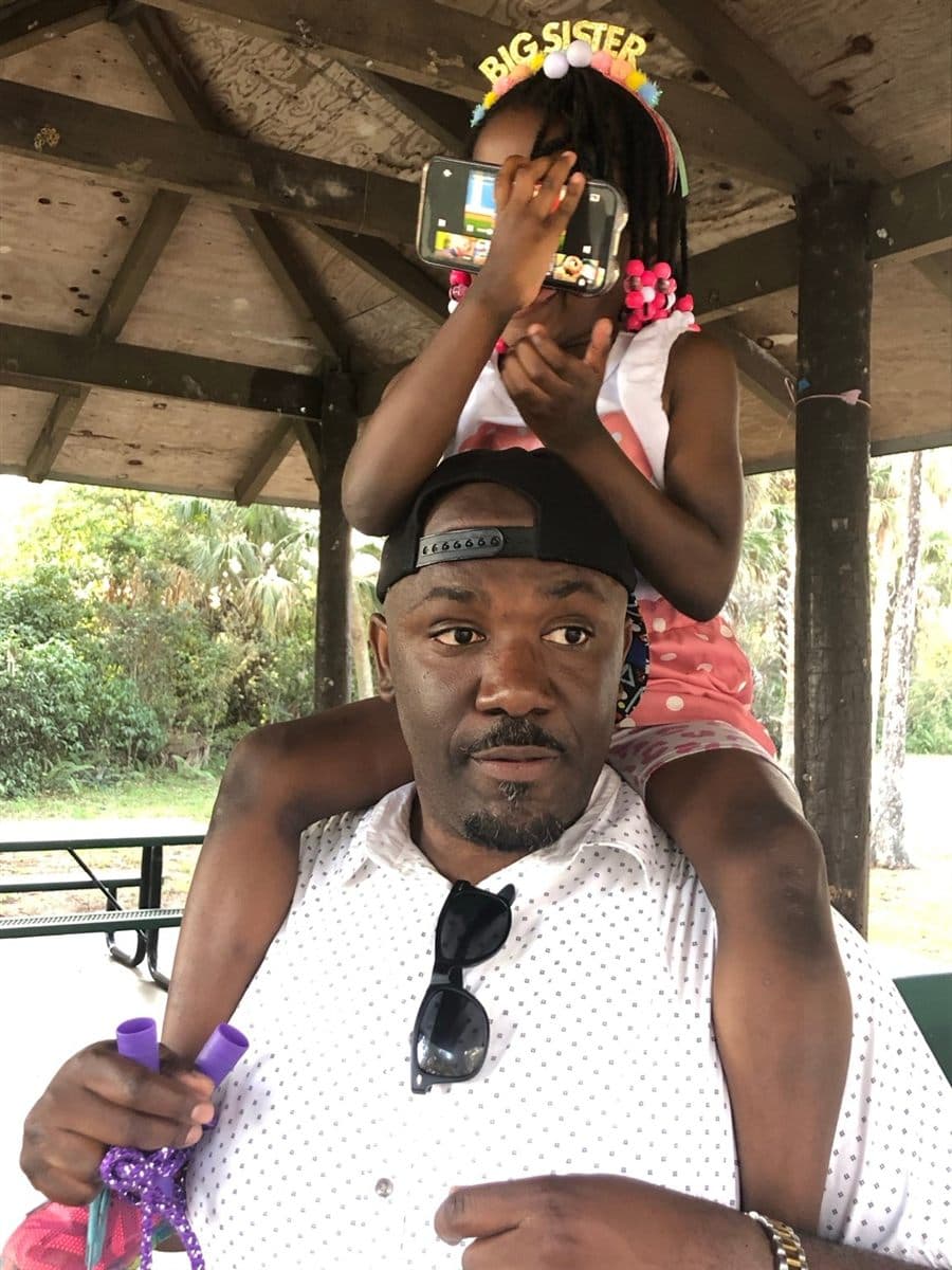 Garaudy Joseph with his daughter sitting on his shoulders at a park